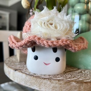 May include: A white ceramic marshmallow figure with a smiling face, wearing a crocheted pink hat adorned with a white fabric flower. The marshmallow is on a decorative wooden stand, with other Easter decorations in the background.