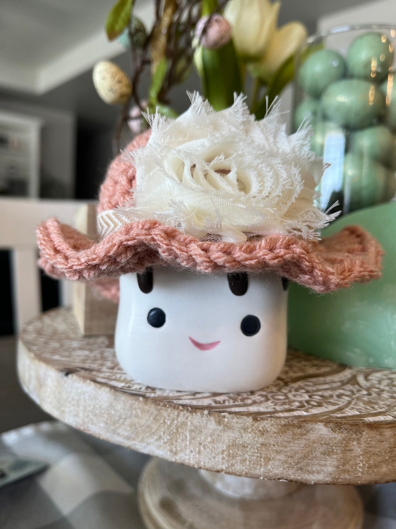 May include: A white ceramic marshmallow figure with a smiling face, wearing a crocheted pink hat adorned with a white fabric flower. The marshmallow is on a decorative wooden stand, with other Easter decorations in the background.