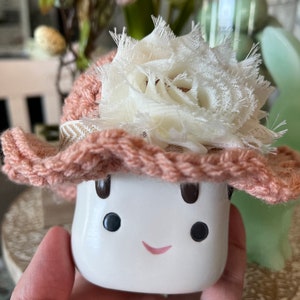 May include: A white ceramic mushroom figurine with a smiling face, black eyes, and a pink mouth. It wears a crocheted peach-colored hat with a cream-colored fabric flower and a patterned band. The figurine is held in a hand.