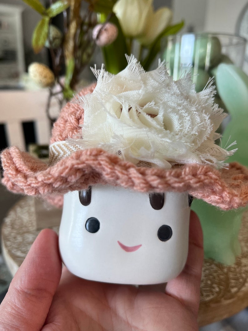 May include: A white ceramic mushroom figurine with a smiling face, black eyes, and a pink mouth. It wears a crocheted peach-colored hat with a cream-colored fabric flower and a patterned band. The figurine is held in a hand.