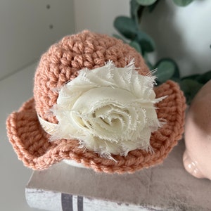 May include: A peach-colored, crocheted hat with a wide brim, adorned with a large, cream-colored fabric flower. The hat has a delicate, handmade appearance, perfect for a baby or child. The flower adds a touch of elegance to the simple design.