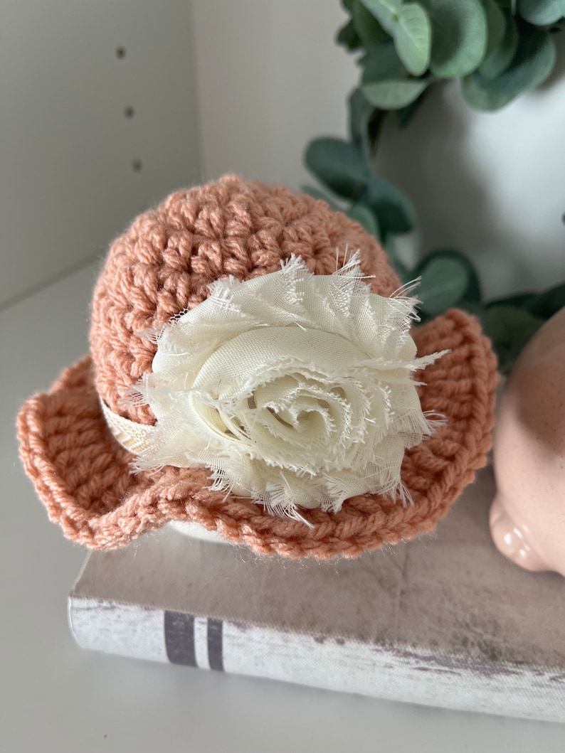 May include: A peach-colored, crocheted hat with a wide brim, adorned with a large, cream-colored fabric flower. The hat has a delicate, handmade appearance, perfect for a baby or child. The flower adds a touch of elegance to the simple design.