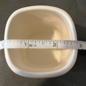 May include: Cream-colored, square ceramic bowl with rounded corners. A measuring tape shows the bowl is approximately 3 inches wide. The bowl is empty and sits on a dark speckled surface.