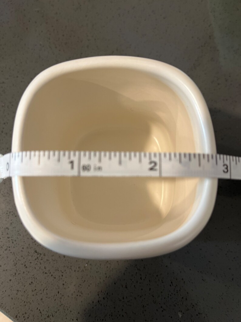 May include: Cream-colored, square ceramic bowl with rounded corners. A measuring tape shows the bowl is approximately 3 inches wide. The bowl is empty and sits on a dark speckled surface.