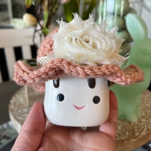 May include: A white ceramic mushroom figurine with a smiling face, wearing a pink crocheted hat adorned with a cream-colored fabric flower. The figurine is held in a hand, with a blurred background featuring Easter decorations.