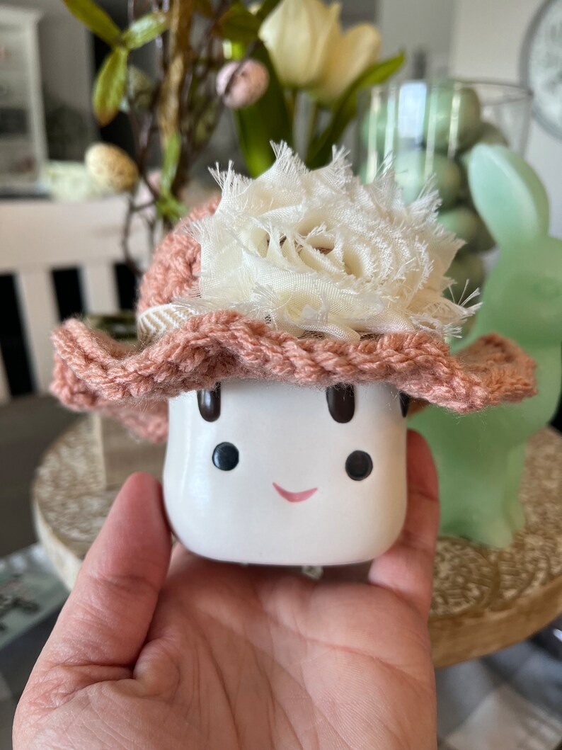 May include: A white ceramic mushroom figurine with a smiling face, wearing a pink crocheted hat adorned with a cream-colored fabric flower. The figurine is held in a hand, with a blurred background featuring Easter decorations.