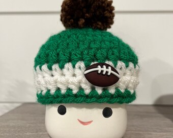 May include: A white ceramic marshmallow figure with a green and white crocheted hat, a brown pom-pom, and a football embellishment. The marshmallow has black eyes and a smiling red mouth. The figure is on a wooden surface.