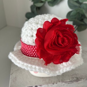 May include: A white crocheted hat with a red fabric flower and a red and white polka dot ribbon. The hat has a wide brim and is sitting on a decorative surface. The hat is a handmade item.