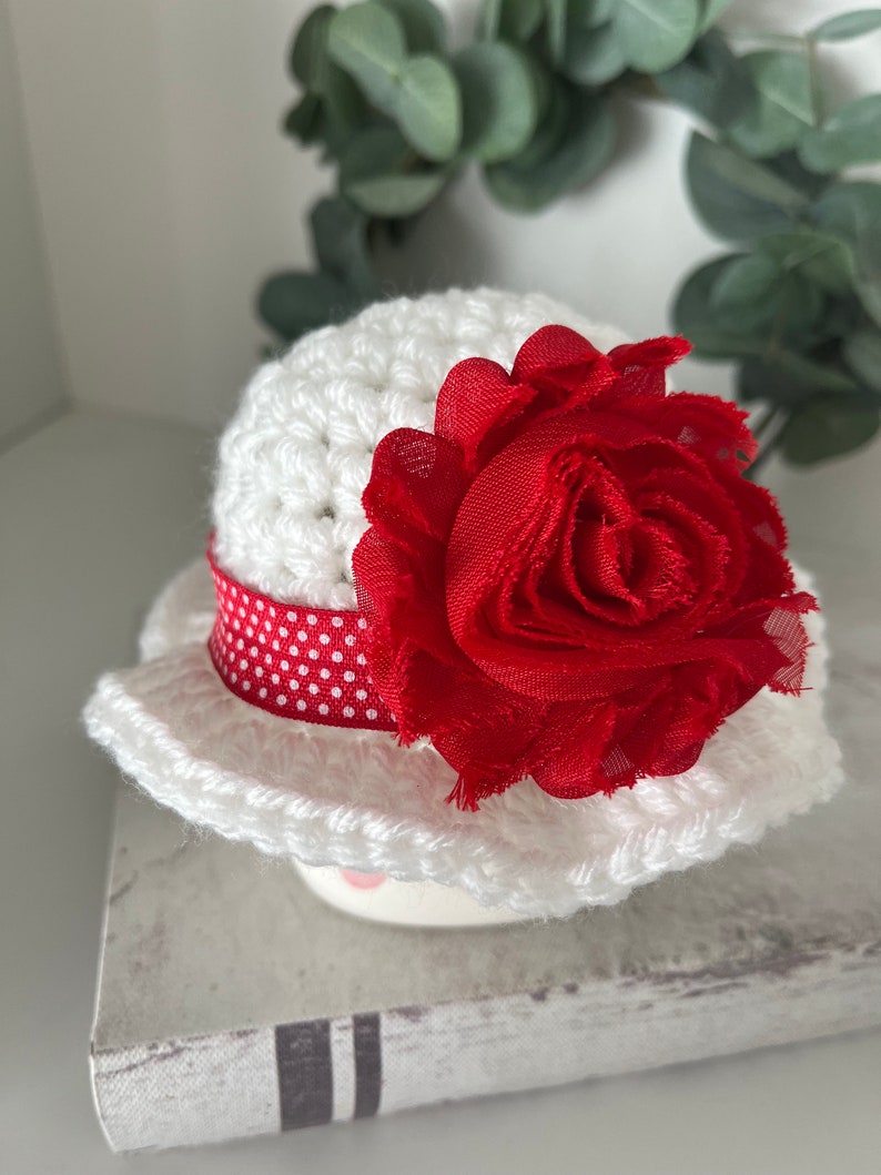 May include: A white crocheted hat with a red fabric flower and a red and white polka dot ribbon. The hat has a wide brim and is sitting on a decorative surface. The hat is a handmade item.