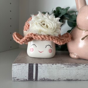 May include: A white ceramic marshmallow-shaped figurine with a smiling face, wearing a crocheted pink hat with a white fabric flower. The figurine is on a decorative book, next to a pink ceramic bunny.