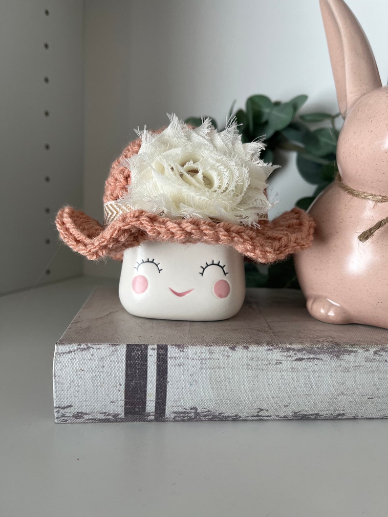 May include: A white ceramic marshmallow-shaped figurine with a smiling face, wearing a crocheted pink hat with a white fabric flower. The figurine is on a decorative book, next to a pink ceramic bunny.