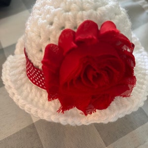 May include: A white crocheted hat with a red fabric flower and a red and white polka dot ribbon. The hat has a rounded crown and a wide brim. The red flower is large and prominent, adding a decorative touch.