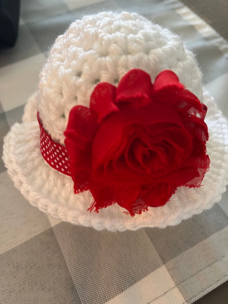 May include: A white crocheted hat with a red fabric flower and a red and white polka dot ribbon. The hat has a rounded crown and a wide brim. The red flower is large and prominent, adding a decorative touch.