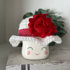 May include: A white ceramic marshmallow-shaped figurine with a smiling face, eyelashes, and rosy cheeks, wearing a white crocheted hat with a red polka-dot ribbon and a large red fabric rose. The figurine is on a decorative book.