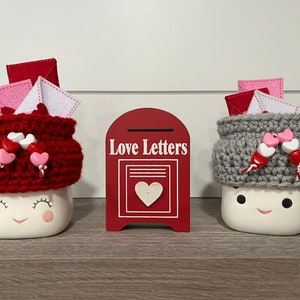 May include: Two ceramic mugs with smiling faces, topped with crocheted hats and felt letters, and heart-shaped embellishments. A red mailbox with "Love Letters" text is between them. The mugs are on a wooden shelf.
