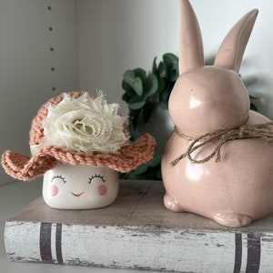 May include: A ceramic bunny figurine and a smiling marshmallow-shaped container wearing a crocheted hat with a white flower. The bunny is light pink with a twine bow. The hat is peach-colored with a white flower. The objects are on a decorative book.