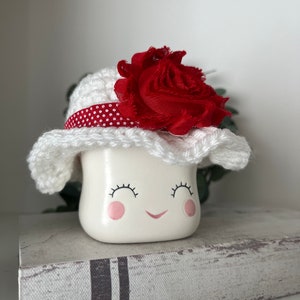 May include: A white ceramic figurine with a smiling face, wearing a white crocheted hat with a red polka dot band and a large red fabric flower. The figurine is sitting on a decorative book.