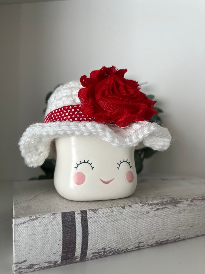 May include: A white ceramic figurine with a smiling face, wearing a white crocheted hat with a red polka dot band and a large red fabric flower. The figurine is sitting on a decorative book.