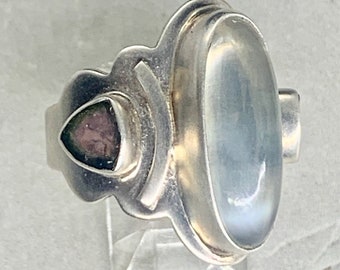 May include: A silver ring with a large oval-shaped clear gemstone and a smaller triangular pink gemstone. The ring has a unique, intricate design.