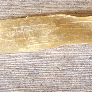 May include: A long, thin, beige object with a slightly wavy texture, possibly a dried leaf or plant part, lies on a weathered, gray wooden surface. The object tapers to a point at one end.