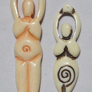 May include: Two cream-colored carved wooden pendants depicting the female form. The larger pendant has a spiral design on the abdomen. The smaller pendant has a spiral design on the lower abdomen and a small carved detail at the bottom.