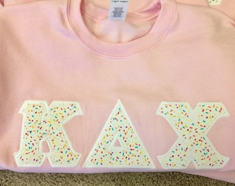 May include: A light pink crewneck sweatshirt featuring large, block-style letters K, A, and X appliqued across the chest. The letters are white with a colourful, multi-coloured sprinkle pattern and are outlined with a thick, white stitched border.