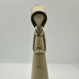 May include: A ceramic figurine of a person wearing a bonnet and a long dress. The figurine is standing with its hands clasped in front of it. The figurine is made of a light brown clay and has a speckled finish.
