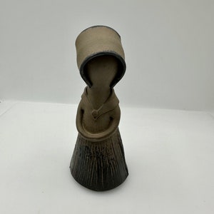 Peut inclure: Une petite figurine en céramique représentant une personne vêtue d'une cape à capuche brune. La figurine est debout, les bras croisés, et a une base texturée de couleur brun foncé.