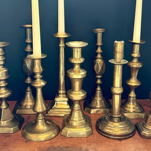 May include: A collection of eight antique brass candle holders with varying designs and heights. The candle holders are arranged on a wooden surface.