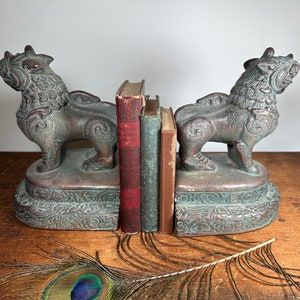 May include: A pair of decorative bookends made of green and brown stone, shaped like mythical lions. The bookends are holding up two books with red and green covers.