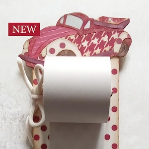 May include: A decorative toilet paper holder with a red and white polka dot pattern. The top features a car-shaped design with a houndstooth pattern. The word "NEW" is displayed on the top left. The holder has a white rope and a red ribbon.