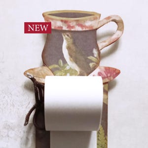 Puede incluir: Un portarrollos de papel decorativo con un diseño floral y de pájaros. La parte superior presenta una forma de taza con una ilustración de pájaro. El soporte tiene una cinta rosa con detalles en forma de corazón y la palabra "NEW" en un rectángulo rojo.