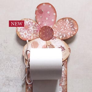Può includere: Un portarotolo di carta decorativo a forma di fiore. Il fiore ha petali rosa e marroni con disegni a motivi. Il supporto ha una base rettangolare con motivi floreali e un accento rosso. La parola "NEW" è in un rettangolo rosso.
