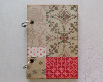Op de afbeelding: Een klein, bruin, met stof bekleed notitieboekje met een patchwork-ontwerp. De omslag heeft verschillende patronen, waaronder bloemen, geometrische vormen en stippen in tinten rood, groen en wit. Het notitieboekje heeft twee zilveren ringen voor de binding.