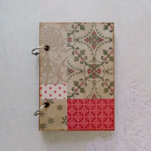 Puede incluir: Un pequeño cuaderno marrón cubierto de tela con un diseño de patchwork. La portada presenta una variedad de patrones, incluyendo diseños florales, geométricos y de lunares en tonos de rojo, verde y blanco. El cuaderno tiene dos anillos plateados para la encuadernación.