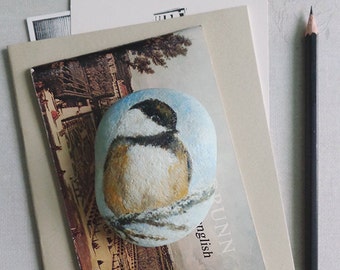 May include: A hand-painted rock with a depiction of a small bird perched on a branch. The rock is sitting on top of a book with the word "RUNN" and "nglish" visible on the cover.