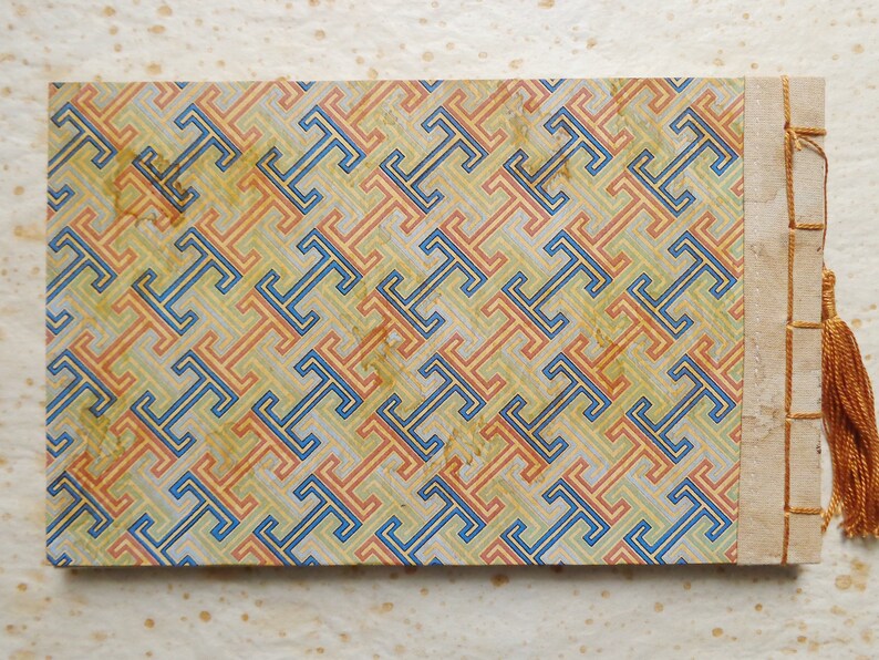 May include: A vintage, hand-bound book with a geometric pattern cover in shades of blue, orange, and yellow. The cover is made of paper and has a decorative binding with a tassel.