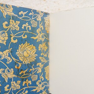 May include: A vintage book with a blue cover featuring a gold floral pattern. The book is open to a blank page.