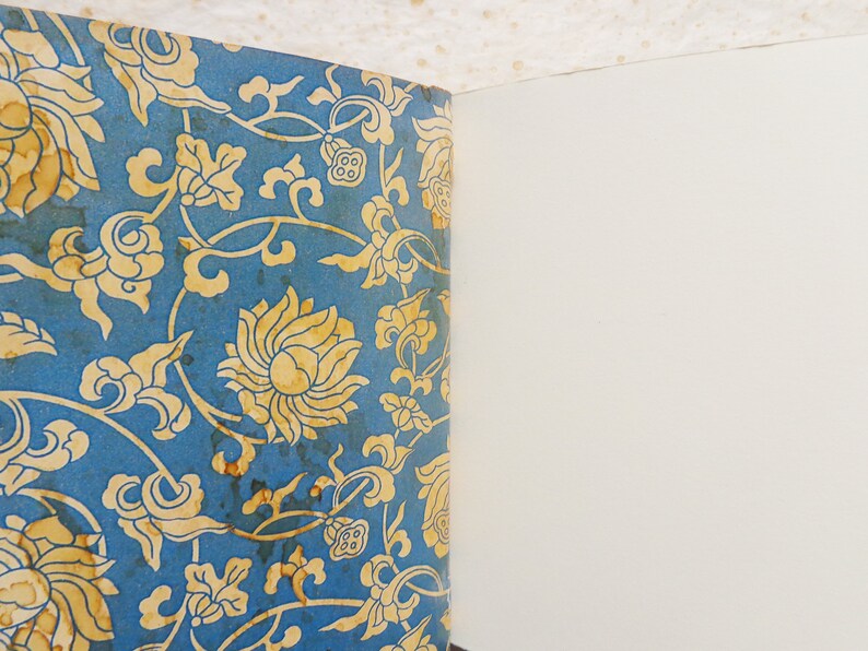 May include: A vintage book with a blue cover featuring a gold floral pattern. The book is open to a blank page.