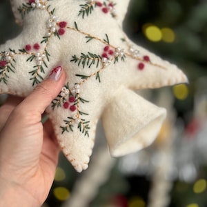May include: Cream-colored felt star ornament with embroidered sprigs of green leaves, red berries, and white beads. The star has a bell-shaped extension. A festive holiday decoration.