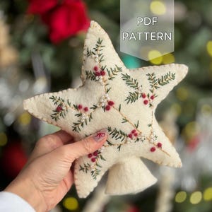 May include: Cream-colored felt star ornament with embroidered green leaves and red berries. The star has a three-dimensional shape and is decorated with small white beads. Text on the image reads "PDF PATTERN".