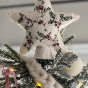 May include: A cream-colored, star-shaped Christmas tree topper. The star is decorated with embroidered green leaves, red berries, and small white beads. It is placed atop a decorated Christmas tree with white ornaments and a pink ribbon.