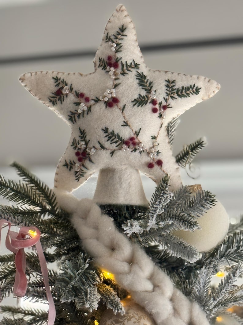 May include: A cream-colored, star-shaped Christmas tree topper. The star is decorated with embroidered green leaves, red berries, and small white beads. It is placed atop a decorated Christmas tree with white ornaments and a pink ribbon.