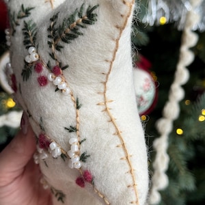 May include: A cream-colored, star-shaped Christmas ornament. The ornament is decorated with embroidered green leaves, red berries, and white beads. The edges are stitched with gold thread. The background includes a Christmas tree and other ornaments.