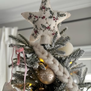 May include: A decorated Christmas tree with a star tree topper. The star is cream-colored with embroidered green leaves and red berries. The tree has white and gold ornaments, a white braided garland, and pink ribbons.