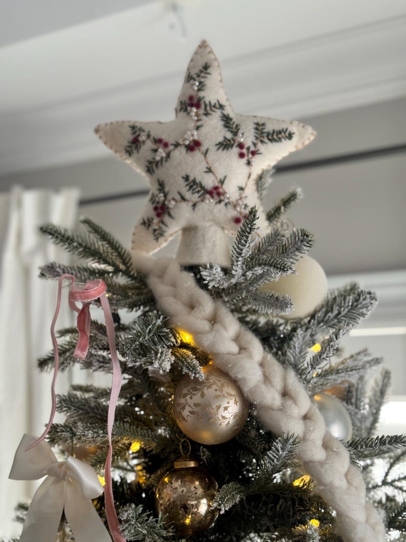 May include: A decorated Christmas tree with a star tree topper. The star is cream-colored with embroidered green leaves and red berries. The tree has white and gold ornaments, a white braided garland, and pink ribbons.