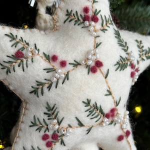 May include: A white felt star Christmas ornament. The star is decorated with green embroidered pine branches, red berry clusters, and small white beads. The ornament is stitched with tan thread and has a loop for hanging.