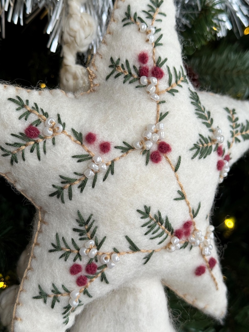 May include: A white felt star Christmas ornament. The star is decorated with green embroidered pine branches, red berry clusters, and small white beads. The ornament is stitched with tan thread and has a loop for hanging.