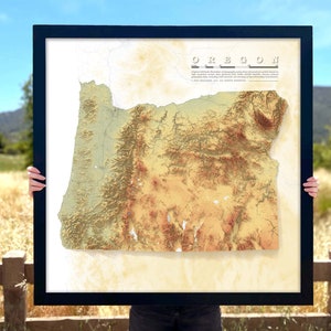 May include: A framed map of Oregon with a topographic relief design. The map is printed on a cream-colored paper with a black frame. The title "OREGON" is printed in black at the top of the map. The map shows the state's major geographic features, including the Cascade Range, the Columbia River, and the Willamette Valley.