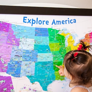 May include: A young child points to a colorful map of the United States with the words "Explore America" at the top. The map is framed in black and hung on a white wall.